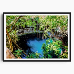 May include: A framed print of a natural sinkhole with vibrant turquoise water. Lush green foliage and hanging vines surround the opening, with sunlight filtering through the trees. The water's surface shows a slight current, creating a dynamic visual.