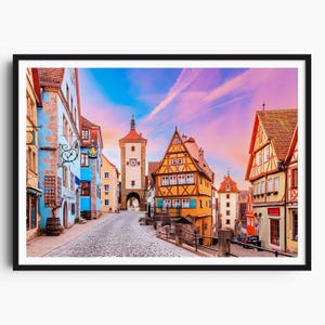 Op de afbeelding: Een ingelijste prent van een Europees stadsplein met kleurrijke gebouwen en een geplaveide straat. De lucht is een mix van blauwe en roze tinten. De gebouwen hebben rode, oranje, gele en blauwe gevels.