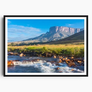 Può includere: Fotografia di paesaggio incorniciata di una catena montuosa con una cima piatta sotto un cielo azzurro. Un fiume scorre in primo piano, con erba verde e rocce lungo le rive. L'immagine è incorniciata da un bordo nero.