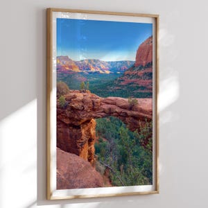 May include: Framed landscape photograph of a natural stone arch bridge in a desert canyon. The image features red rock formations, green vegetation, and a clear blue sky. The photograph is encased in a wooden frame.