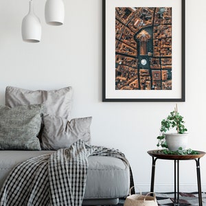 May include: A framed aerial photograph of a city with a large, circular building in the center. The building has a dome-shaped roof and is surrounded by smaller buildings. The photograph is in a black frame and is hanging on a white wall.