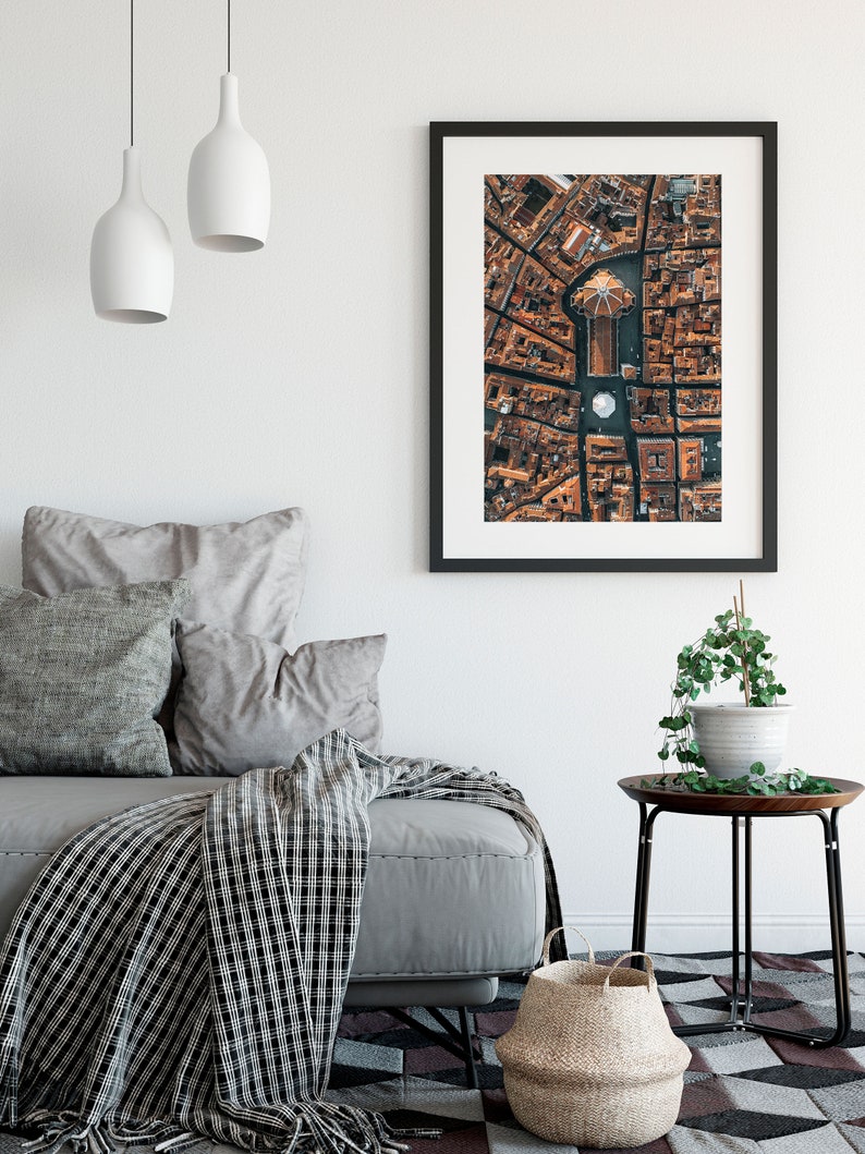 May include: A framed aerial photograph of a city with a large, circular building in the center. The building has a dome-shaped roof and is surrounded by smaller buildings. The photograph is in a black frame and is hanging on a white wall.