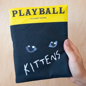 May include: A black fabric item with the word "PLAYBALL" in bold black letters on a yellow band. Below, the item is black with white text reading "KITTENS" and cartoon cat eyes.