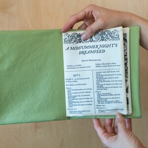 May include: A light green book cover holds open pages of a book titled "A Midsummer Night's Dreamfeed." The text is in black ink with decorative borders. The book is held open by a person.