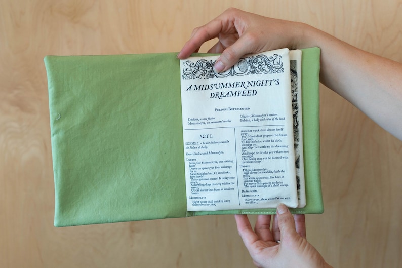 May include: A light green book cover holds open pages of a book titled "A Midsummer Night's Dreamfeed." The text is in black ink with decorative borders. The book is held open by a person.