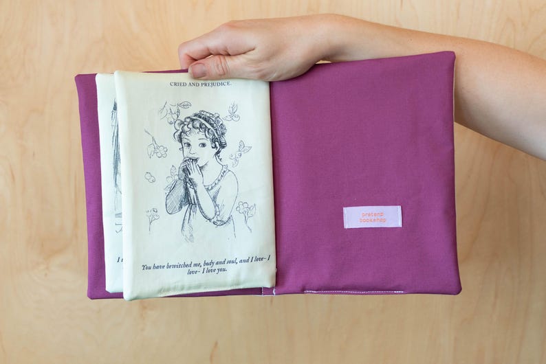 May include: A purple book sleeve holds a vintage book page with a black and white illustration and text from "Pride and Prejudice." The sleeve has a small rectangular label with text. The book is open, revealing the printed pages.
