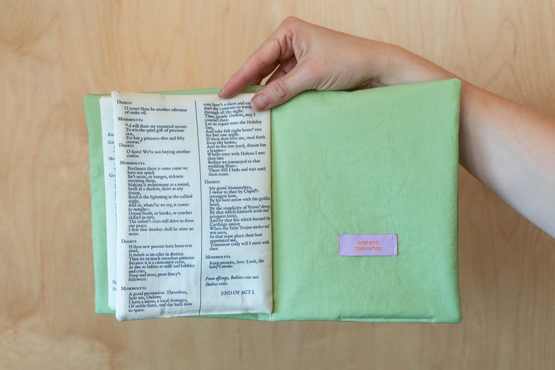 May include: A light green book cover with a book inside, held open by a hand. The book pages are visible, with text from a play. A small, rectangular label with the text "pretend bookshop" is on the cover.