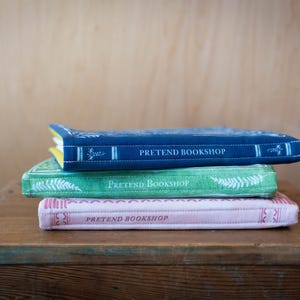 May include: Three stacked, decorative books with the text "PRETEND BOOKSHOP" on the covers. The top book is navy blue, the middle is green, and the bottom is pink. The books are on a wooden surface.