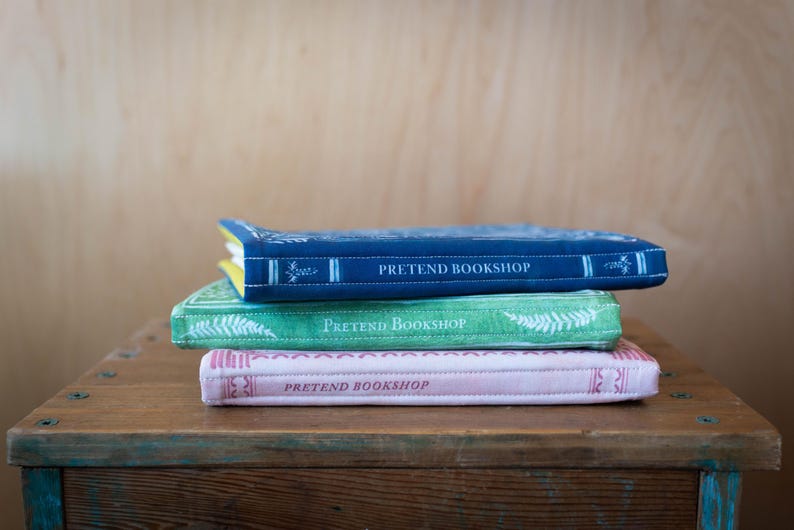 May include: Three stacked, decorative books with the text "PRETEND BOOKSHOP" on the covers. The top book is navy blue, the middle is green, and the bottom is pink. The books are on a wooden surface.