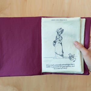 May include: A burgundy fabric book cover with a cream-colored page featuring a pen and ink illustration and text from "Cried and Prejudice". The illustration depicts a woman in a dress and hat with a duck. The text reads "It is a truth universally acknowledged that a toddler in possession of a good appetite, must be in want of a berry."