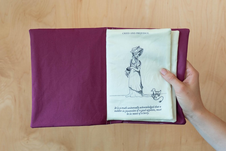May include: A burgundy fabric book cover with a cream-colored page featuring a pen and ink illustration and text from "Cried and Prejudice". The illustration depicts a woman in a dress and hat with a duck. The text reads "It is a truth universally acknowledged that a toddler in possession of a good appetite, must be in want of a berry."