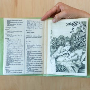 May include: An open book with green fabric covers, displaying printed text and a black and white illustration. The illustration depicts a person tending to another person in a forest setting. The text appears to be from a play.