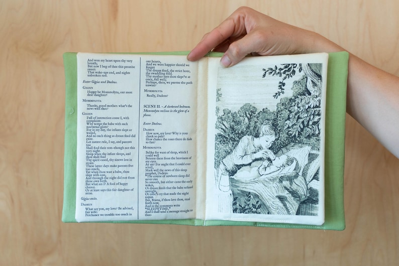 May include: An open book with green fabric covers, displaying printed text and a black and white illustration. The illustration depicts a person tending to another person in a forest setting. The text appears to be from a play.