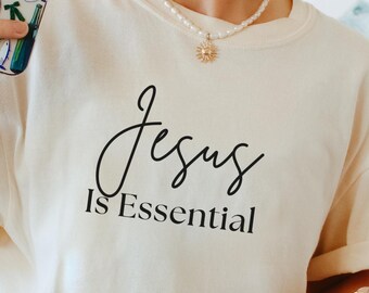 May include: Ivory-colored t-shirt with the text "Jesus Is Essential" in a black script font. The shirt is paired with blue and white striped bottoms. A clear glass with a wooden lid and a straw is held in the hand.