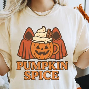 May include: Ivory-colored t-shirt with a pumpkin spice graphic. The design features a smiling jack-o'-lantern mug with whipped cream and cinnamon sticks, flanked by two figures in orange cloaks. The words "PUMPKIN SPICE" are printed below in orange.