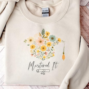Peut inclure: Un sweat-shirt crème avec un motif de chapeau de graduation floral et le texte "Mastered It" avec un diplôme en dessous.