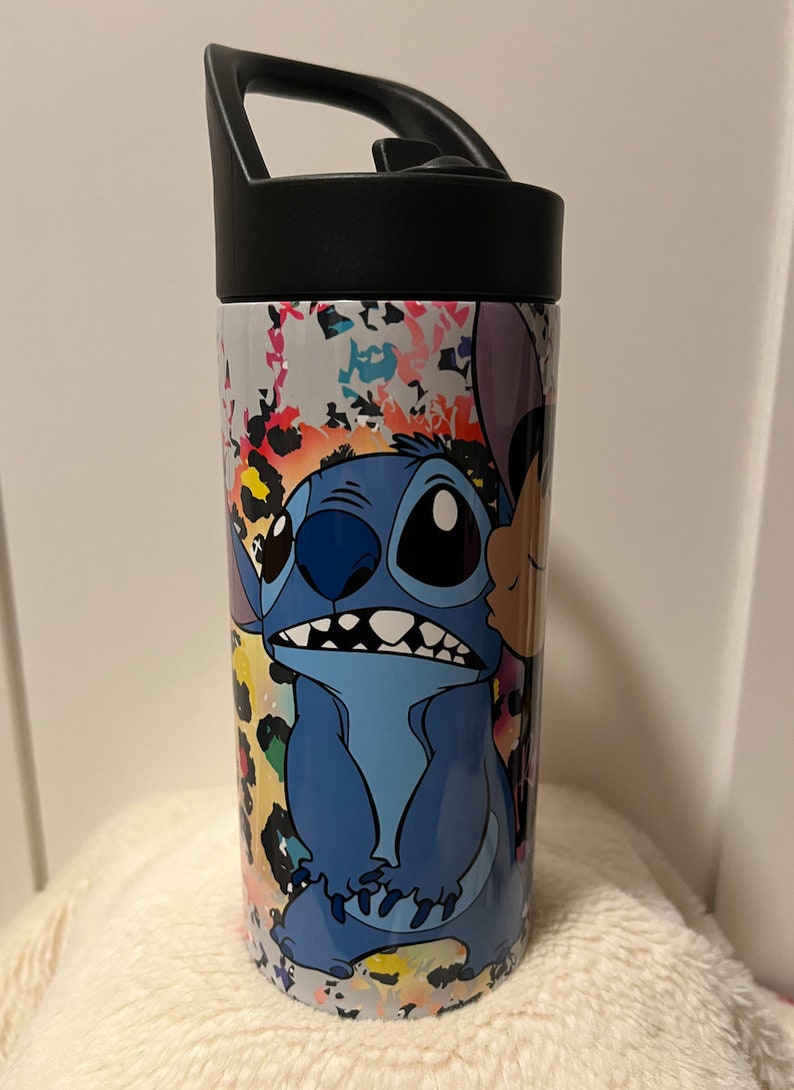 May include: A stainless steel water bottle with a black lid and handle. The bottle features a colorful abstract design with a cartoon image of Stitch from Lilo & Stitch.