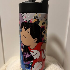 May include: A white stainless steel travel mug with a black lid and handle. The mug features a colorful abstract design with a cartoon image of Lilo and Stitch from the Disney movie Lilo & Stitch. The name "Rylee" is written in purple on the mug.