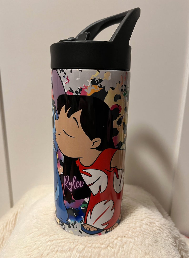 May include: A white stainless steel travel mug with a black lid and handle. The mug features a colorful abstract design with a cartoon image of Lilo and Stitch from the Disney movie Lilo & Stitch. The name "Rylee" is written in purple on the mug.