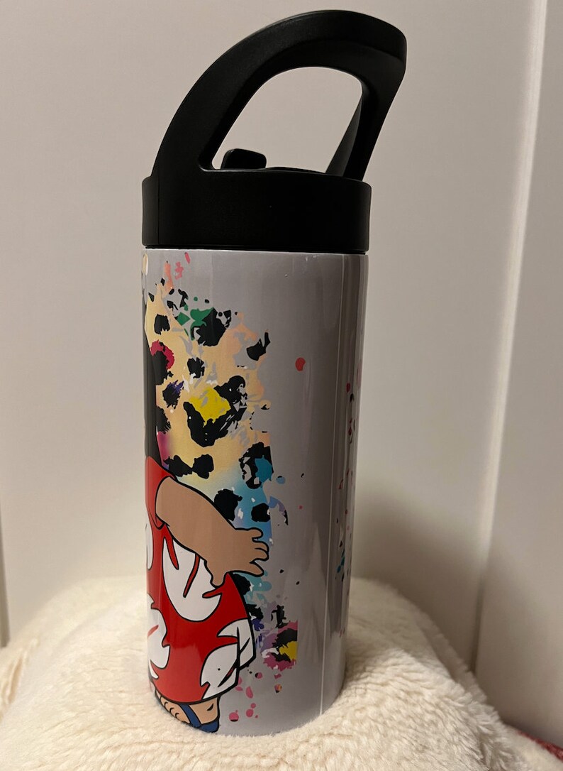 May include: A gray stainless steel water bottle with a black lid and handle. The bottle features a colorful graphic of a cartoon character wearing a red and white floral dress. The background of the graphic is a gray and white splatter pattern.