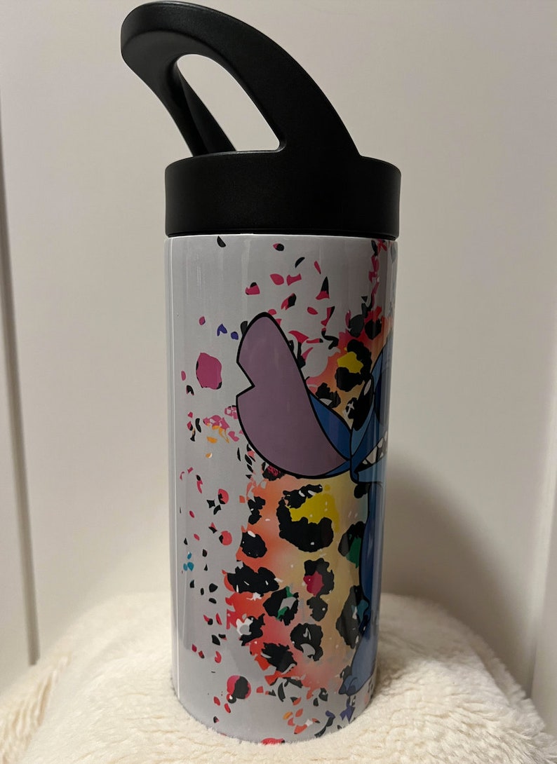May include: A white stainless steel water bottle with a black lid and handle. The bottle has a colorful design featuring a cartoon character, Stitch, from the Disney movie Lilo & Stitch, with a leopard print background.