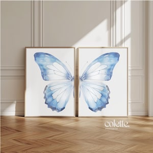 May include: Two framed prints of a watercolor painting of a blue butterfly with white wings. The butterfly is centered in each frame and fills the entire space. The frames are a light wood color.