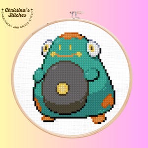 May include: A cross-stitch embroidery hoop featuring a teal, orange, and gray pixelated creature. The creature has large yellow eyes and a circular gray object on its belly. The text "Christina's Stitches" is in the upper left corner.