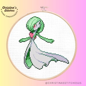 May include: Cross stitch pattern of a green and white Pokemon character, Gardevoir, in a white dress with a pink heart on her chest. The pattern is on a white background and is framed in a wooden hoop.