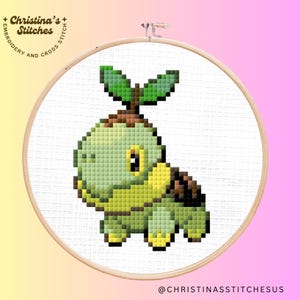 May include: A cross-stitch pattern of a green and brown Pokemon character with a leafy head. The character is on a white background with a wooden embroidery hoop.
