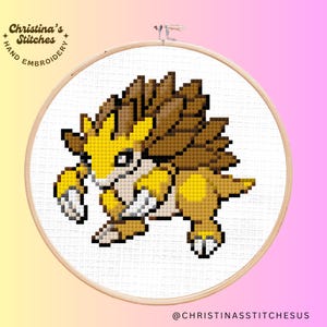 May include: Cross stitch pattern of a yellow and brown Pokemon character with a spiky back. The character is running and has a determined expression. The pattern is on a white background and is framed in a wooden hoop.