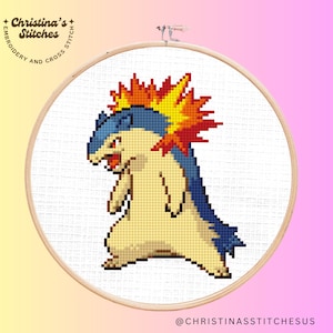 Puede incluir: Bordado de punto de cruz de un personaje Typhlosion en un aro de madera. El personaje es principalmente amarillo, azul y rojo, con un diseño en forma de llama en la cabeza. El aro está sobre un fondo degradado rosa y amarillo. El texto en el aro dice "Christina's Stitches".