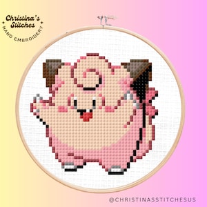 Könnte beinhalten: Ein pinkfarbenes und braunes Pixel-Stickmuster eines Pokemon-Charakters, Jigglypuff, auf weißem Hintergrund. Das Design befindet sich auf einem hölzernen Stickrahmen.