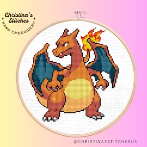 May include: Cross-stitch embroidery pattern of a red and orange Charizard Pokemon with blue wings and a flame tail. The pattern is on white Aida cloth in a wooden embroidery hoop.