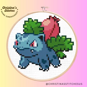 Peut inclure: Une broderie au point de croix représentant un personnage Pokemon bleu et vert avec une fleur rouge sur la tête. Le personnage est entouré d'un fond blanc et d'un cercle de broderie en bois.