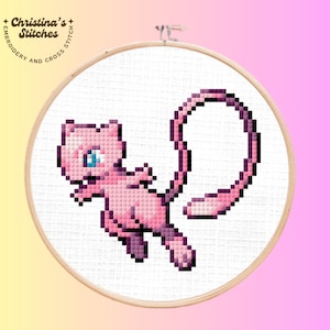 May include: A cross-stitch embroidery hoop featuring a pink pixelated Mew character with blue eyes. The design is set against a white fabric background, framed by a wooden hoop. The text "Christina's Stitches" is visible.