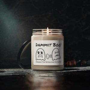 Puede incluir: Una vela encendida en un tarro de cristal con tapa negra. La etiqueta dice "DAMMIT BOO!" con fantasmas de dibujos animados. La vela está sobre una superficie oscura y reflectante, creando un ambiente espeluznante.