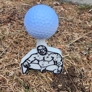 May include: A light blue golf ball sits atop a white tee featuring a black and white illustration of a muscular figure. The tee is designed to hold the golf ball in place during a golf swing.