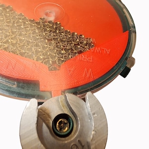 May include: A clear plastic container with an orange interior holds numerous small, brass-colored objects. The container has the text "ALWAYS WIPE PRIMERS" printed on it. A metal tool with a circular opening is positioned below the container.