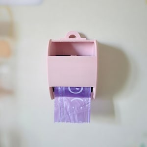 May include: A light pink wall-mounted bag dispenser with a curved top and a small handle. A roll of purple bags with a smiling face design is visible, dispensing from the bottom. The dispenser is mounted on a light green wall.
