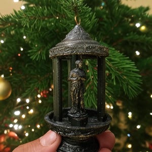 May include: A detailed, dark bronze Christmas ornament featuring a miniature statue within a gazebo-like structure. The ornament has intricate detailing and is suspended by a gold-colored hook. The background is a blurred Christmas tree.