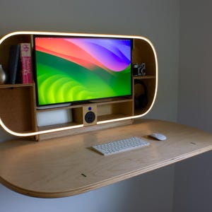 May include: A modern, wall-mounted desk with a built-in monitor and shelves. The desk is made of light-colored wood and features a curved design with a glowing light strip. A keyboard and mouse sit on the desk surface. The monitor displays a colorful abstract image.