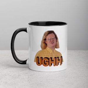 Puede incluir: Taza de cerámica blanca con interior y asa negros. La taza presenta una persona con gafas y la palabra "UGHH" en letras naranjas y marrones. La persona lleva una camisa amarilla a cuadros.