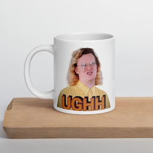 Puede incluir: Taza de cerámica blanca con una persona con gafas y una camisa a cuadros amarilla. La taza tiene la palabra "UGHH" en letras naranjas y marrones. La taza está sobre una superficie de madera.