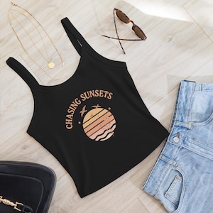 Puede incluir: Camiseta sin mangas negra con el texto "CHASING SUNSETS" y un gráfico de puesta de sol. El gráfico presenta un círculo con rayas naranjas y rosas, pájaros y plantas. También se ven gafas de sol y un collar dorado.