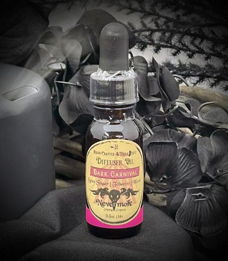 May include: A dark brown glass bottle of "Dark Carnival" diffuser oil with a black dropper. The label is yellow with pink accents and features the text "Nevermore Company". The bottle contains 0.5 fl oz.