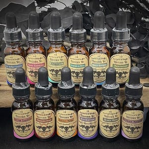 May include: A collection of brown glass diffuser oil bottles with black droppers. Each bottle has a label with a unique name, such as "Dark Carnival" and "Victorian Mourning." The labels feature a skull and crossbones design. The bottles are arranged on a wooden surface.