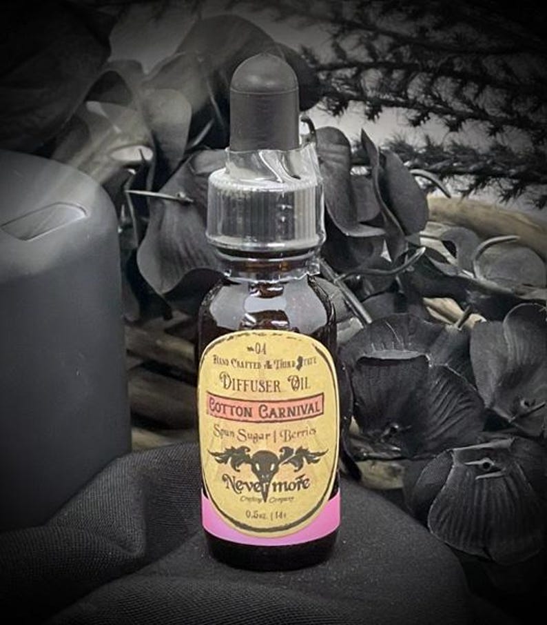 May include: A small, dark glass bottle of diffuser oil with a black dropper. The label reads "Cotton Carnival" and "Nevermore". The label is yellow with pink accents and features a skull and floral design. The bottle contains 0.5 oz (14 ml) of oil.