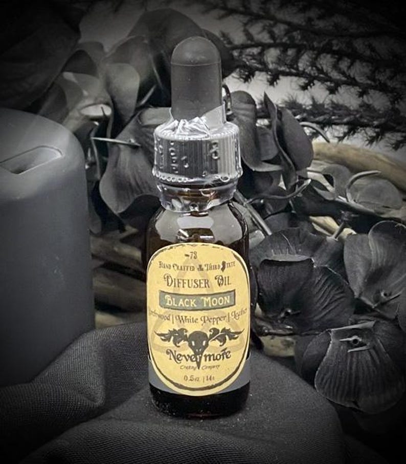 May include: A dark brown glass diffuser oil bottle with a black dropper. The label reads "Black Moon" and "Nevermore". The bottle contains 0.5 oz / 14 ml of oil. The background includes black flowers and a black candle.