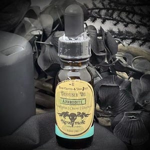 May include: A small, brown glass bottle of Aphrodite diffuser oil with a black dropper. The label reads "Nevermore" and lists ingredients. The bottle is 0.5 fl oz. The background includes dark foliage and a black candle.