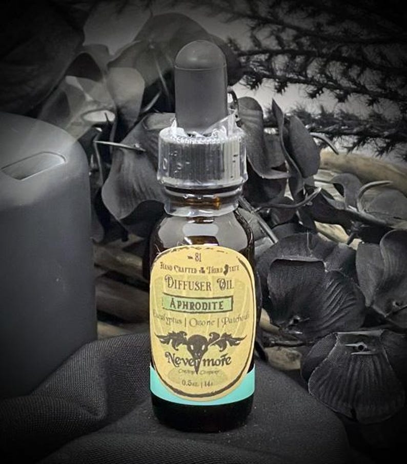 May include: A small, brown glass bottle of Aphrodite diffuser oil with a black dropper. The label reads "Nevermore" and lists ingredients. The bottle is 0.5 fl oz. The background includes dark foliage and a black candle.